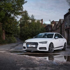 Front three-quarter view of a white Audi sedan parked in a moody urban alley with brick walls and overgrown vegetation