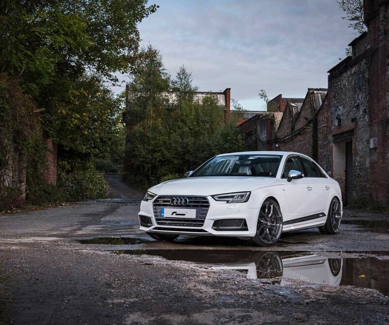 Front three-quarter view of a white Audi sedan parked in a moody urban alley with brick walls and overgrown vegetation