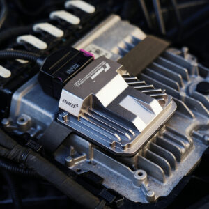 VWR030000-C8RS6_6-1jpg - OPSHOLDERS Close-up of a metallic automotive electronic module mounted on an engine, with cooling fins and a black connector cable nearby.