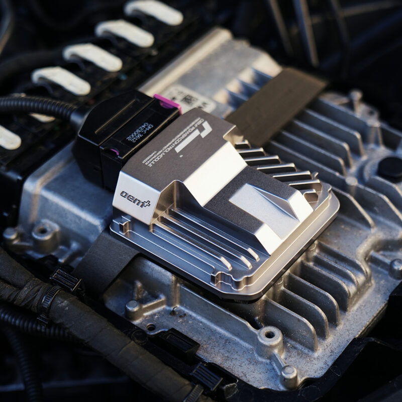 VWR030000-C8RS6_6-1jpg - OPSHOLDERS Close-up of a metallic automotive electronic module mounted on an engine, with cooling fins and a black connector cable nearby.