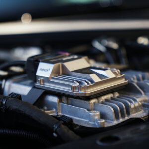 VWR030000-C8RS6_7-1jpg - OPSHOLDERS Close-up of a silver automotive electronic module mounted on a car engine, with hoses nearby.