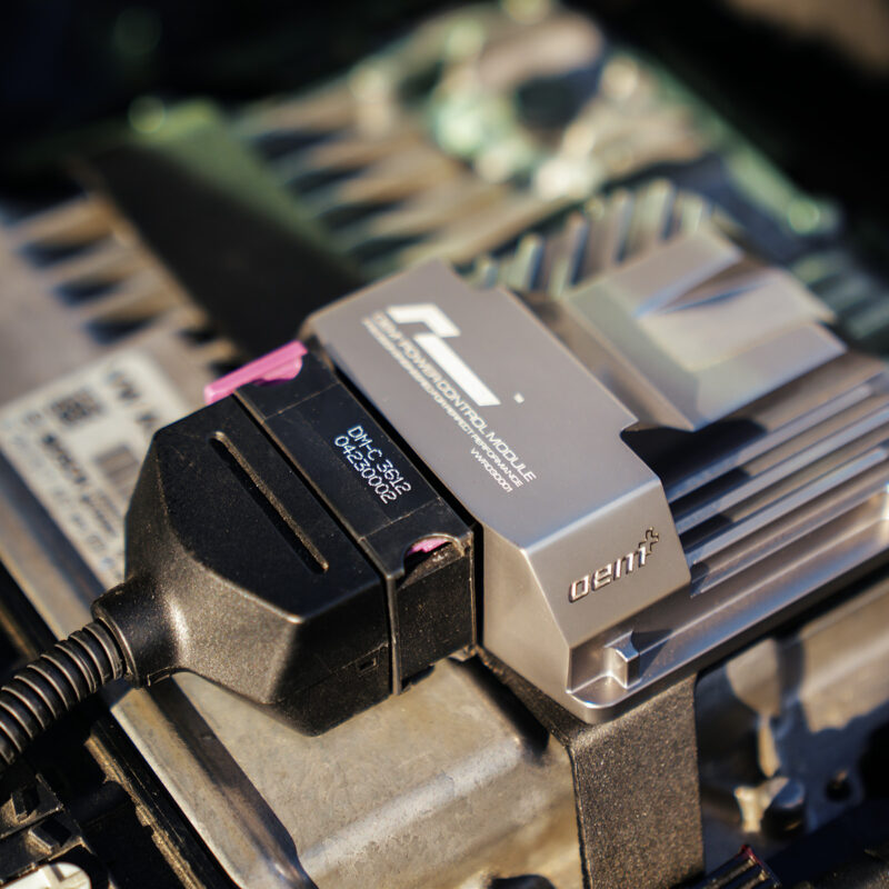VWR030000-C8RS6_8-1jpg - OPSHOLDERS Close-up of an automotive OBD diagnostic module plugged into a vehicle's diagnostic port with a black connector nearby.