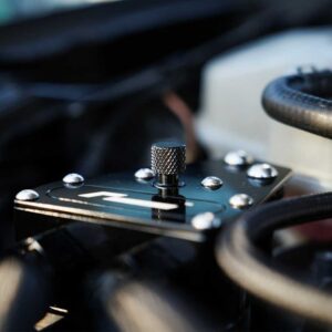 Close-up of a knurled metal knob on a black control plate with screws, inside a car engine bay.