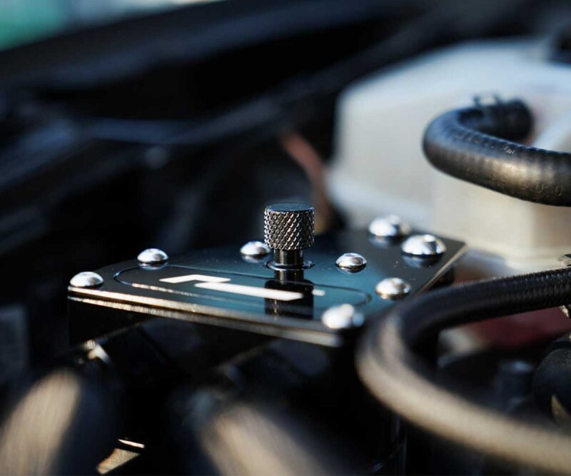 Close-up of a knurled metal knob on a black control plate with screws, inside a car engine bay.