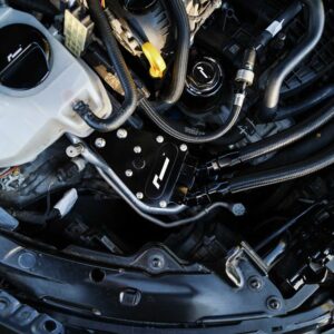 Close-up of a car engine bay showing braided hoses, a black mounting plate with bolts, and surrounding components.