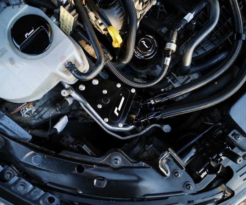 Close-up of a car engine bay showing braided hoses, a black mounting plate with bolts, and surrounding components.