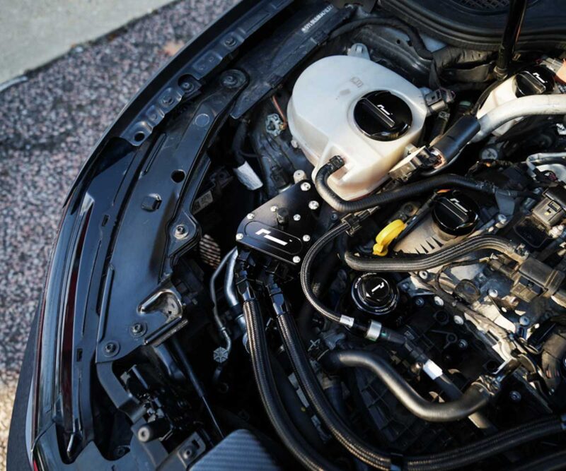 Close-up of a car engine bay showing an aftermarket aluminum strut brace and various hoses and fittings to the left and center of the image