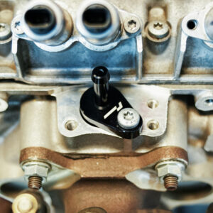 VWR140014_7-1jpg - OPSHOLDERS Close-up of a car engine's valve train: a black adjustment lever mounted on the cylinder head with bolts visible around it.