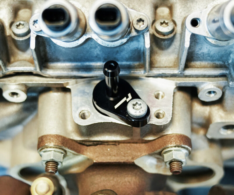 VWR140014_7-1jpg - OPSHOLDERS Close-up of a car engine's valve train: a black adjustment lever mounted on the cylinder head with bolts visible around it.