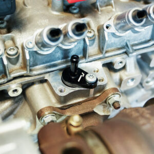 VWR140014_9jpg - OPSHOLDERS Close-up of a car engine cylinder head with fuel injector ports and a small black valve/lever mounted on the metal housing.