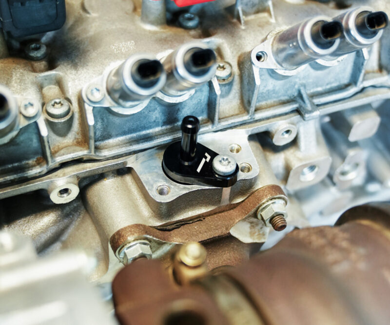VWR140014_9jpg - OPSHOLDERS Close-up of a car engine cylinder head with fuel injector ports and a small black valve/lever mounted on the metal housing.