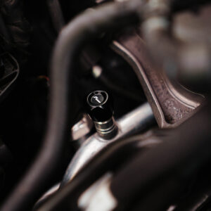 Close-up of a car engine bay showing a black cap with a white arrow, surrounded by hoses and metal brackets.