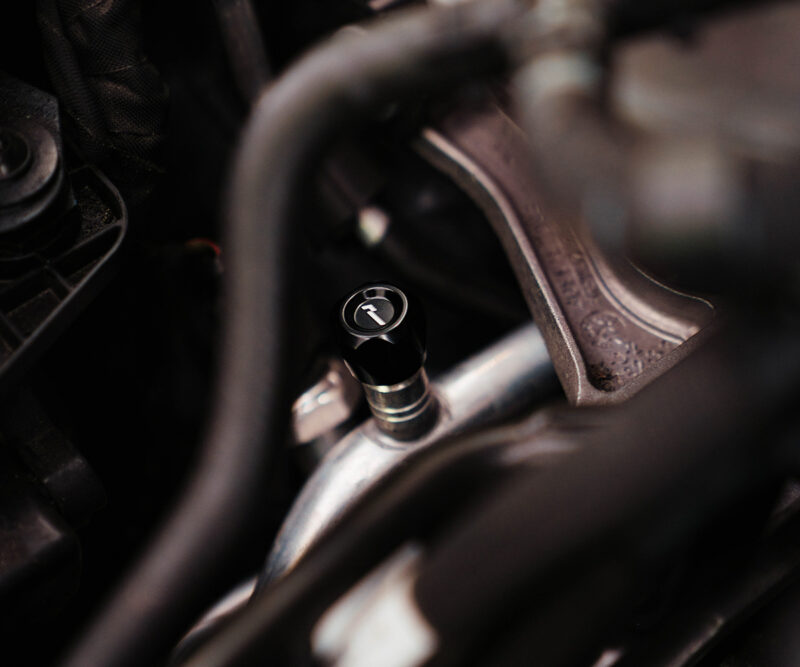Close-up of a car engine bay showing a black cap with a white arrow, surrounded by hoses and metal brackets.