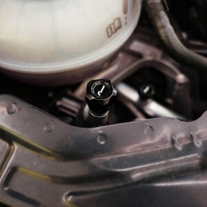 Close-up of a car engine bay featuring a black oil cap with a white symbol, surrounded by hoses and metal framework.
