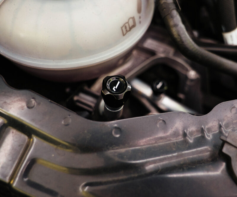 Close-up of a car engine bay featuring a black oil cap with a white symbol, surrounded by hoses and metal framework.