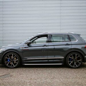 Side view of a dark gray luxury SUV with large black multi-spoke wheels parked beside a corrugated metal wall on pavement.