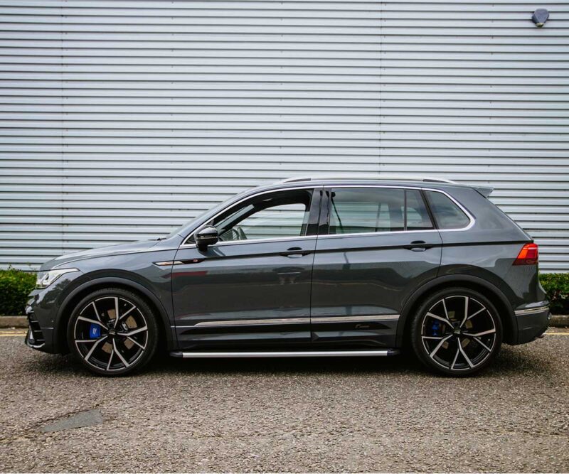 Side view of a dark gray luxury SUV with large black multi-spoke wheels parked beside a corrugated metal wall on pavement.