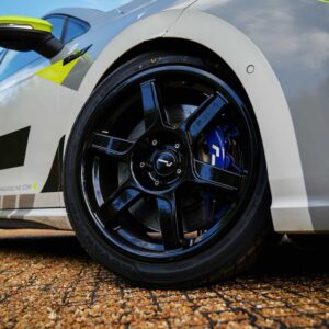 Close-up of a black alloy wheel and blue brake caliper on a white race car, with a gravel surface nearby.