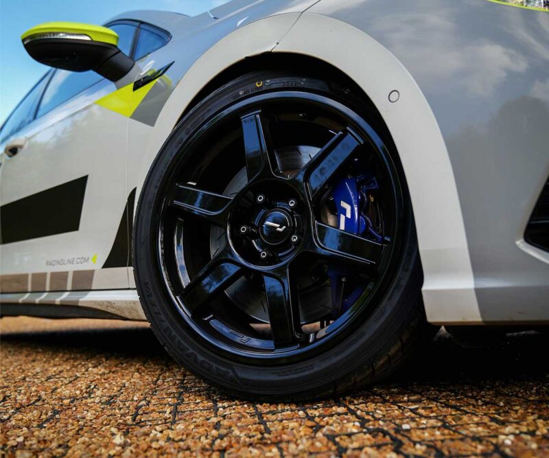 Close-up of a black alloy wheel and blue brake caliper on a white race car, with a gravel surface nearby.