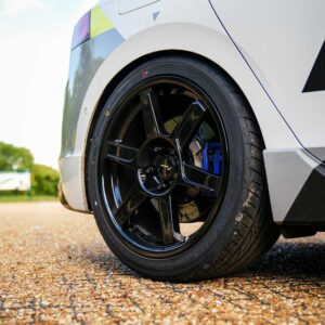 Close-up of a glossy black alloy wheel with a blue brake caliper on a white car, parked on gravel.