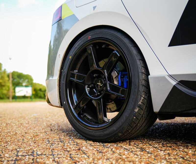Close-up of a glossy black alloy wheel with a blue brake caliper on a white car, parked on gravel.