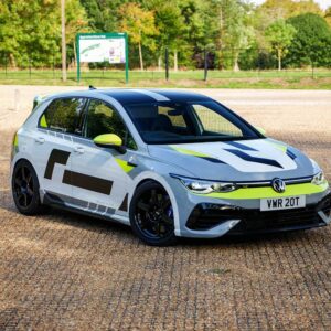White VW hatchback race car with neon yellow-green accents parked on a gravel surface, rear-ready spoiler visible. Plate: VWR 20T.