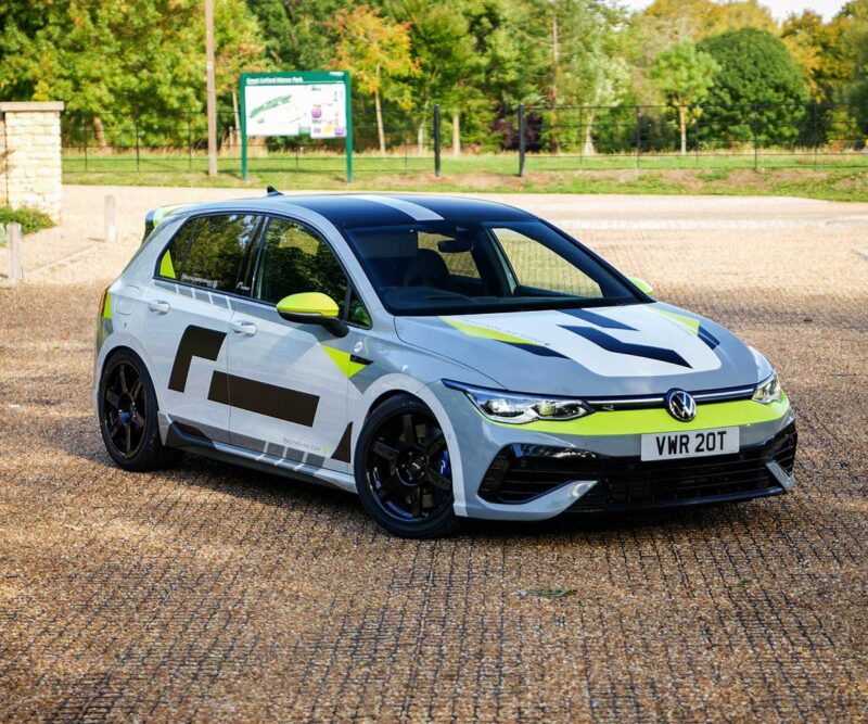 White VW hatchback race car with neon yellow-green accents parked on a gravel surface, rear-ready spoiler visible. Plate: VWR 20T.
