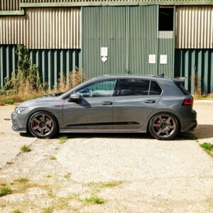 VWR610401_14jpg - OPSHOLDERS Side view of a gray hatchback with aftermarket wheels parked on a gravel lot in front of a green corrugated metal industrial building.