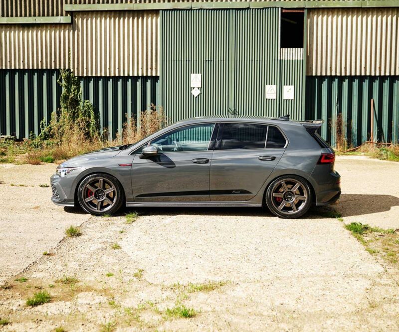 VWR610401_14jpg - OPSHOLDERS Side view of a gray hatchback with aftermarket wheels parked on a gravel lot in front of a green corrugated metal industrial building.