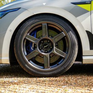 VWR610401_5jpg - OPSHOLDERS Close-up of a car wheel: alloy rim, blue brake caliper, and brake disc visible on a high-visibility gray vehicle with yellow graphics, parked on gravel.