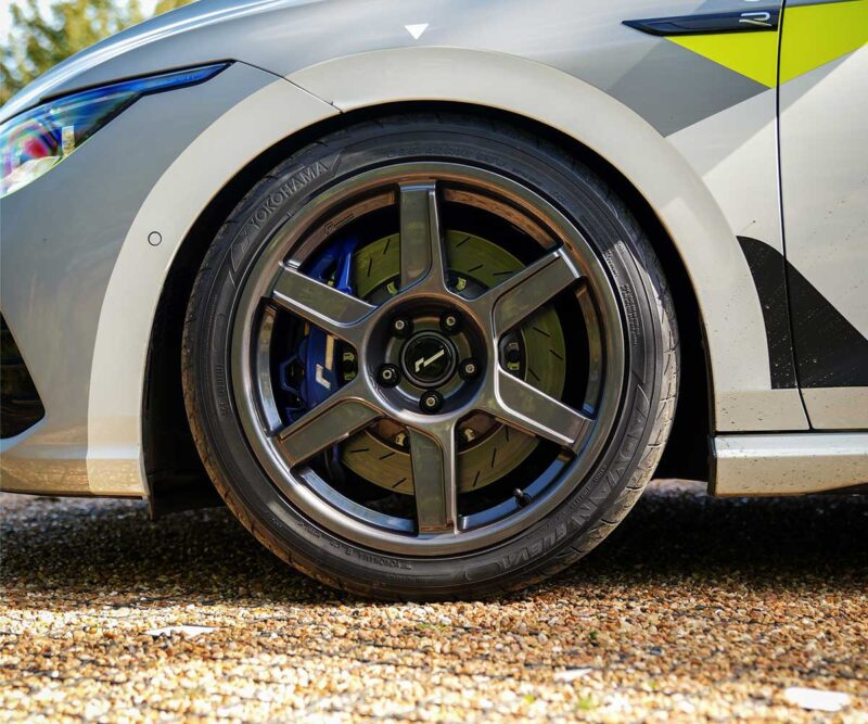 VWR610401_5jpg - OPSHOLDERS Close-up of a car wheel: alloy rim, blue brake caliper, and brake disc visible on a high-visibility gray vehicle with yellow graphics, parked on gravel.