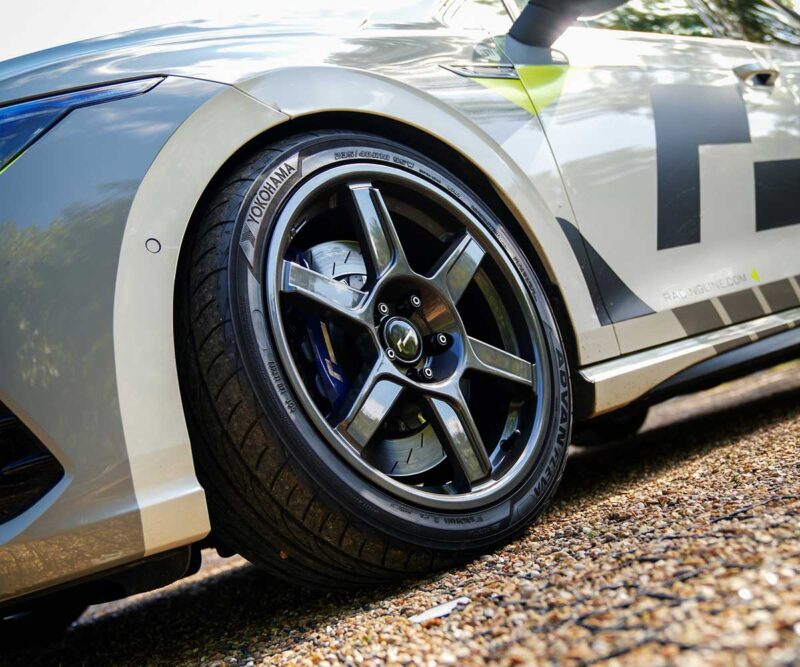 VWR610401_6jpg - OPSHOLDERS Close-up of a race car wheel with a Yokohama tire and alloy rim, parked on gravel, part of a white/gray race car body with large graphics.