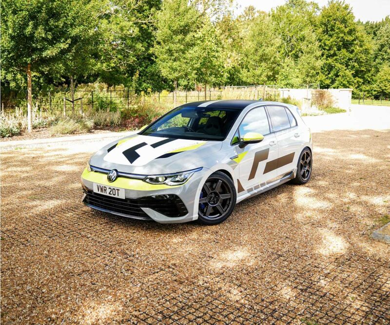 VWR610401_7jpg - OPSHOLDERS White hatchback with yellow and gray racing graphics parked on a gravel driveway with green trees in the background.
