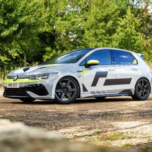 VWR610401_8jpg - OPSHOLDERS White Volkswagen hatchback with black and neon yellow racing decals parked on a gravel lot, trees in the background