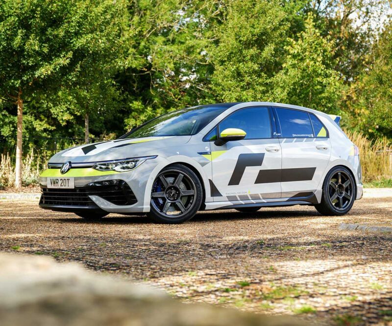 VWR610401_8jpg - OPSHOLDERS White Volkswagen hatchback with black and neon yellow racing decals parked on a gravel lot, trees in the background