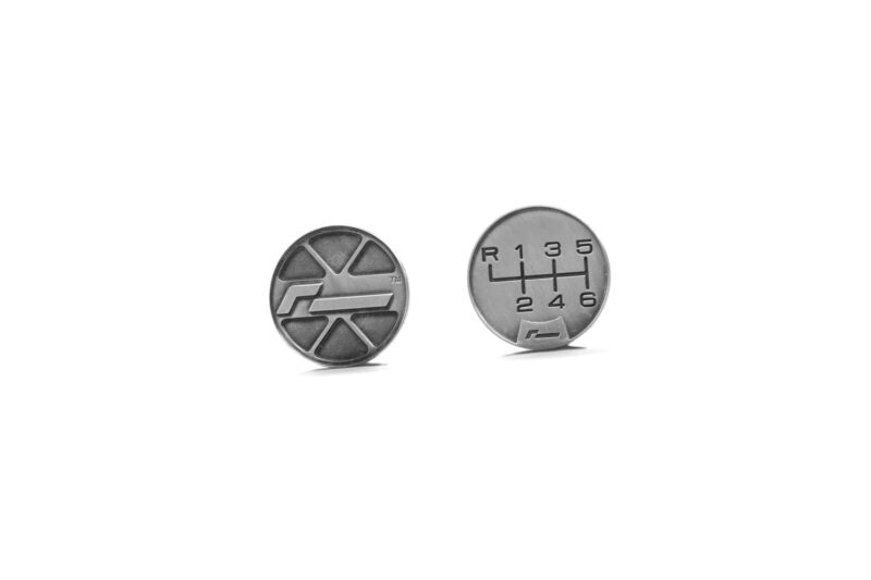 Two silver coins on a white background; left coin has a geometric logo, right coin shows a car gearshift diagram with R and numbers 1–6.