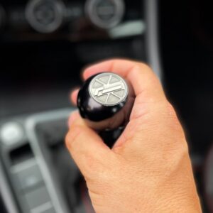 Close-up of a hand gripping a car's gear shifter with a circular metal emblem on top, inside a blurred vehicle interior. (informative)