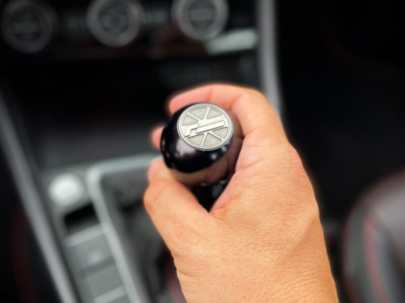 Close-up of a hand gripping a car's gear shifter with a circular metal emblem on top, inside a blurred vehicle interior. (informative)