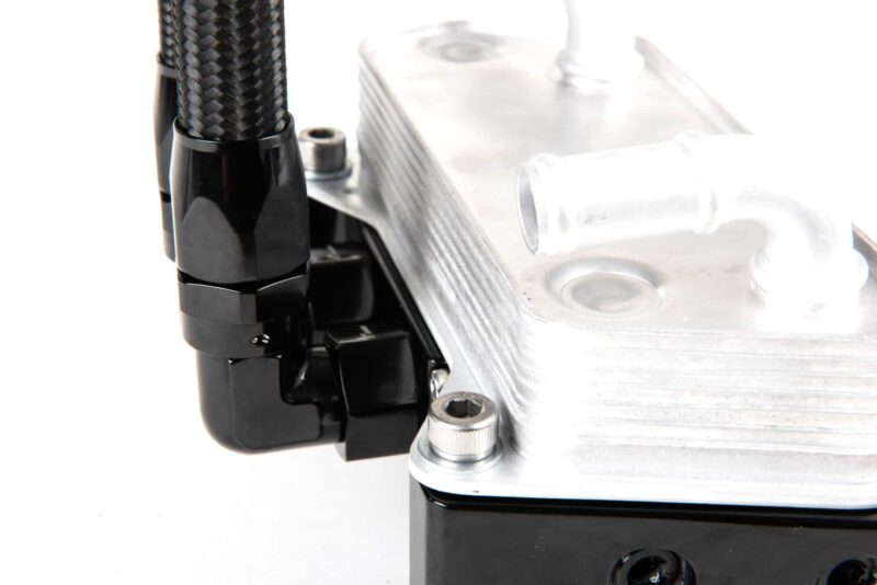 Macro close-up of a braided hose connected to a black mounting bracket beside a silver machined metal block with bolts.