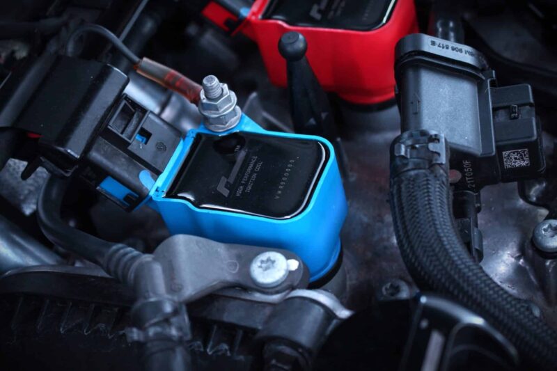 Close-up of a car battery terminal: blue terminal cover with a red jumper cable attached to a metal post under the hood.