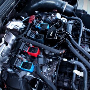 Close-up of a car engine bay with wiring, hoses and blue/red aftermarket electronic modules.
