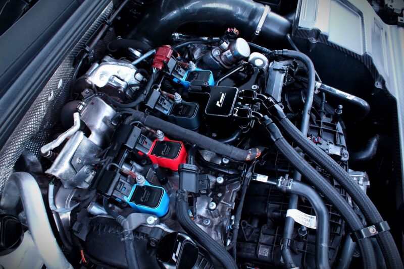 Close-up of a car engine bay with wiring, hoses and blue/red aftermarket electronic modules.