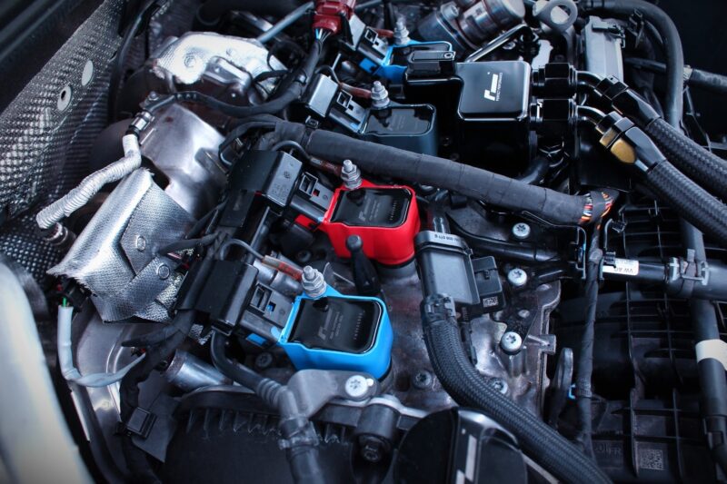 Engine bay with assorted electronic diagnostic modules (red, blue) plugged into wiring harnesses amid hoses and metal components.