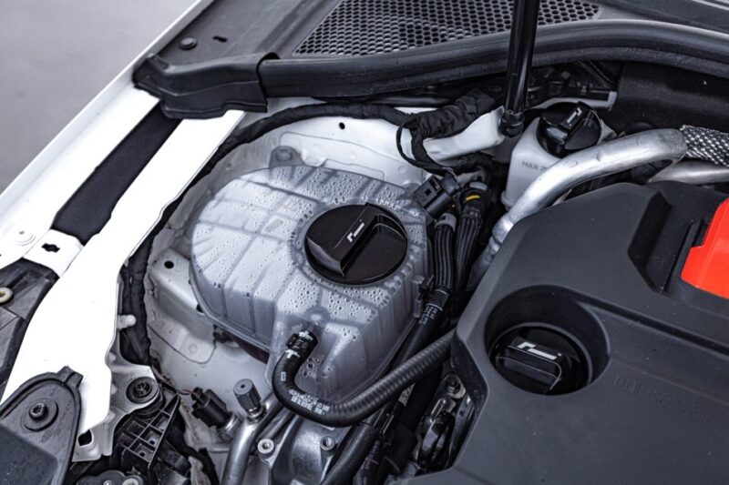 oil-cap-gen4-mqb-evo_51745577247_o-1jpg - OPSHOLDERS Close-up of a car engine bay showing the translucent coolant reservoir with a black cap and surrounding hoses and components.