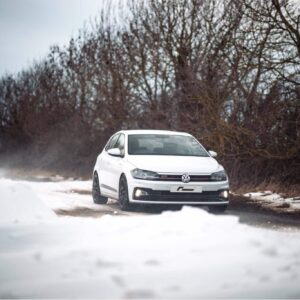 polo-gti-aw-intercooler_53248438804_o-scaled-2jpg - OPSHOLDERS White Volkswagen sedan driving on a snow-covered road with bare trees in the background.