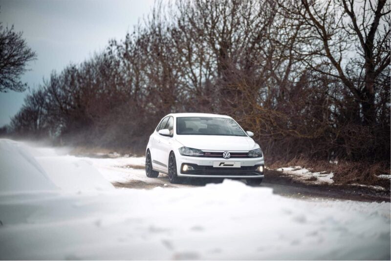 polo-gti-aw-intercooler_53248438804_o-scaled-2jpg - OPSHOLDERS White Volkswagen sedan driving on a snow-covered road with bare trees in the background.