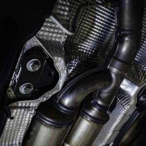 propshaft-bearing-alignment-kit_51768771773_o-scaled-2jpg - OPSHOLDERS Close-up of a metallic exhaust pipe and mounting bracket with carbon-fiber texture in a dark setting, likely part of a vehicle engine bay.
