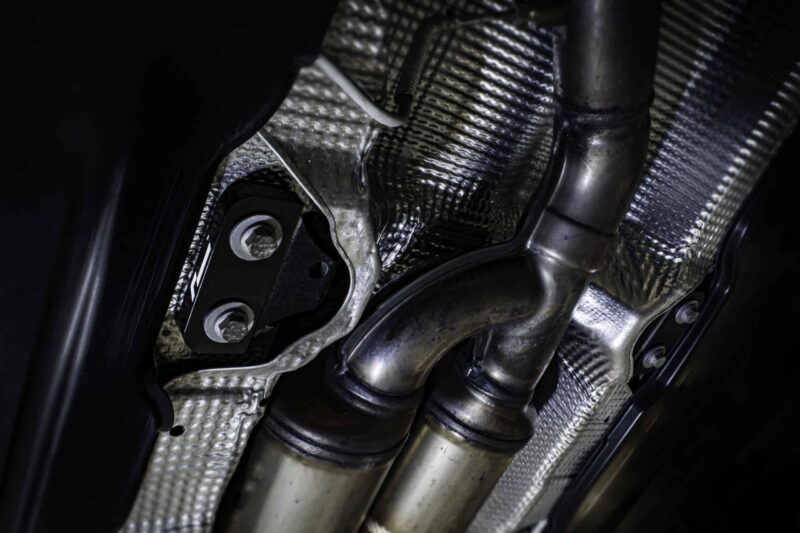 propshaft-bearing-alignment-kit_51768771773_o-scaled-2jpg - OPSHOLDERS Close-up of a metallic exhaust pipe and mounting bracket with carbon-fiber texture in a dark setting, likely part of a vehicle engine bay.