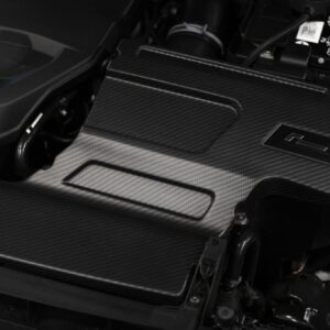 Close-up of a carbon-fiber car interior surface with a small rectangular metal cover and visible wiring in the background.
