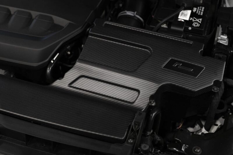 Close-up of a carbon-fiber car interior surface with a small rectangular metal cover and visible wiring in the background.